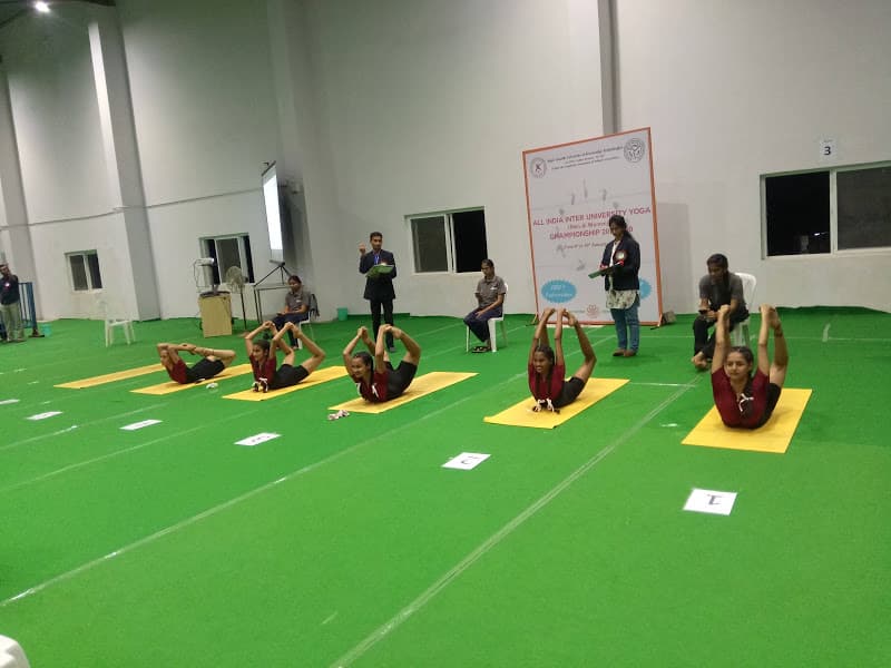 Five young women holding aligned Dhanurasana (Bow Pose) on yellow mats during inter-university yoga championship competition